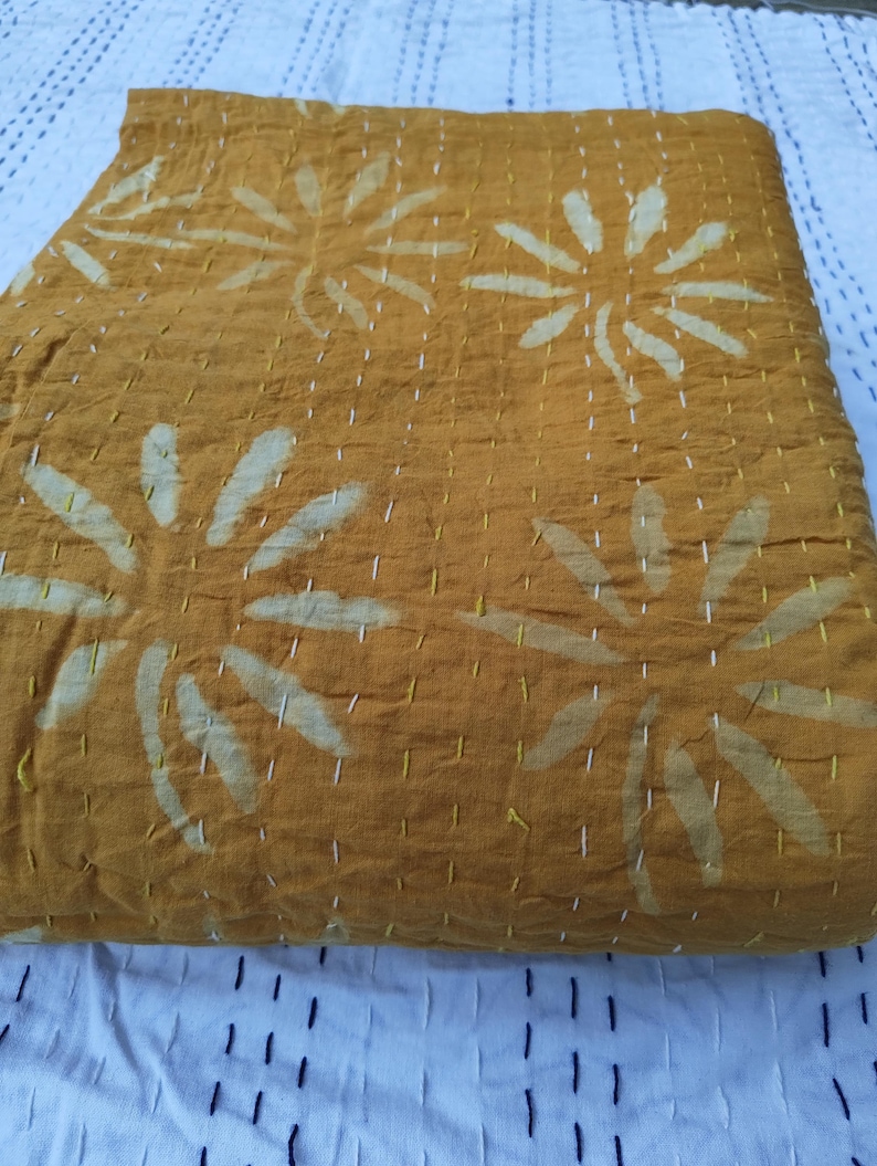 May include: Folded mustard-colored textile with large, white floral patterns. The fabric is stitched with white thread, creating a textured appearance. The textile is resting on a white surface with a blue and black striped pattern.