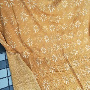 May include: Mustard-colored textile with a repeating white floral pattern. The fabric appears to be quilted, with visible stitching. The reverse side of the textile is a solid mustard color. The textile is folded, showing both sides.