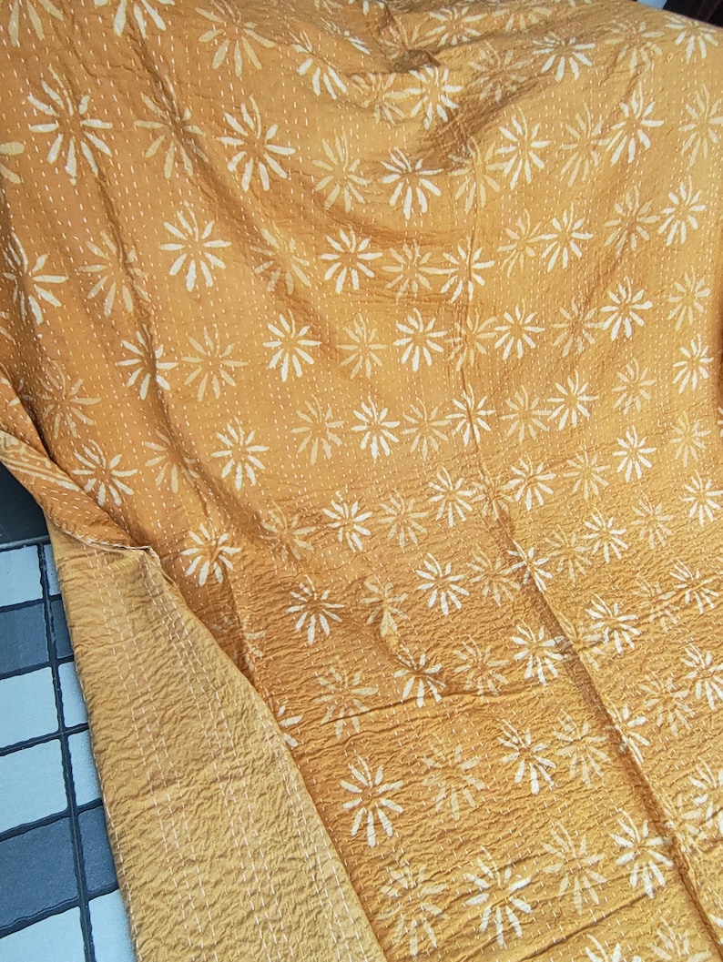 May include: Mustard-colored textile with a repeating white floral pattern. The fabric appears to be quilted, with visible stitching. The reverse side of the textile is a solid mustard color. The textile is folded, showing both sides.