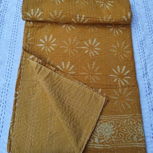 May include: Mustard yellow kantha quilt with white floral and leaf patterns. The quilt is folded and partially unfolded, showcasing the intricate stitching and design. The edges feature a decorative border.
