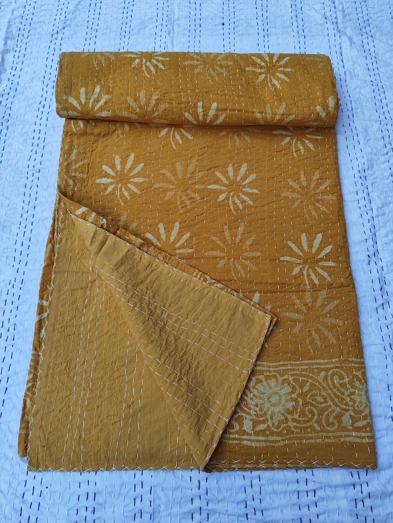 May include: Mustard yellow kantha quilt with white floral and leaf patterns. The quilt is folded and partially unfolded, showcasing the intricate stitching and design. The edges feature a decorative border.