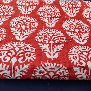 May include: A red and white patterned quilt. The quilt features a repeating design of stylized floral motifs in white against a red background. The stitching is visible, adding texture to the fabric. The quilt is folded.