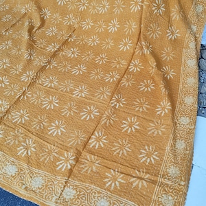 May include: Mustard-colored textile with a repeating white floral pattern. The fabric has a textured appearance, with a decorative border featuring a floral and scroll design. The overall aesthetic suggests a handcrafted or vintage item.