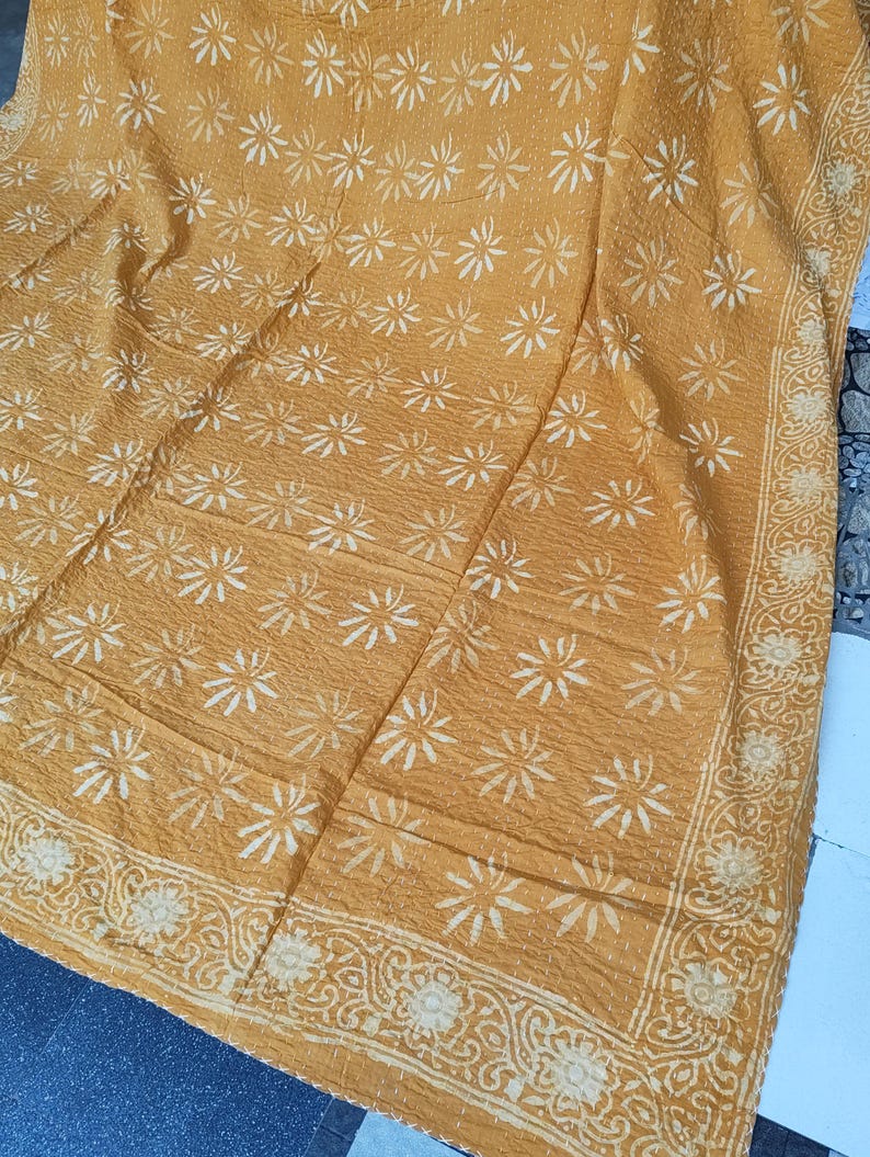 May include: Mustard-colored textile with a repeating white floral pattern. The fabric has a textured appearance, with a decorative border featuring a floral and scroll design. The overall aesthetic suggests a handcrafted or vintage item.