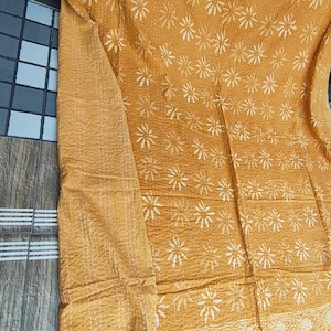 May include: Mustard-colored textile with a repeating white floral pattern. The fabric has a quilted texture with visible stitching. A detailed border with floral designs is visible along one edge. The textile is likely a blanket or throw.
