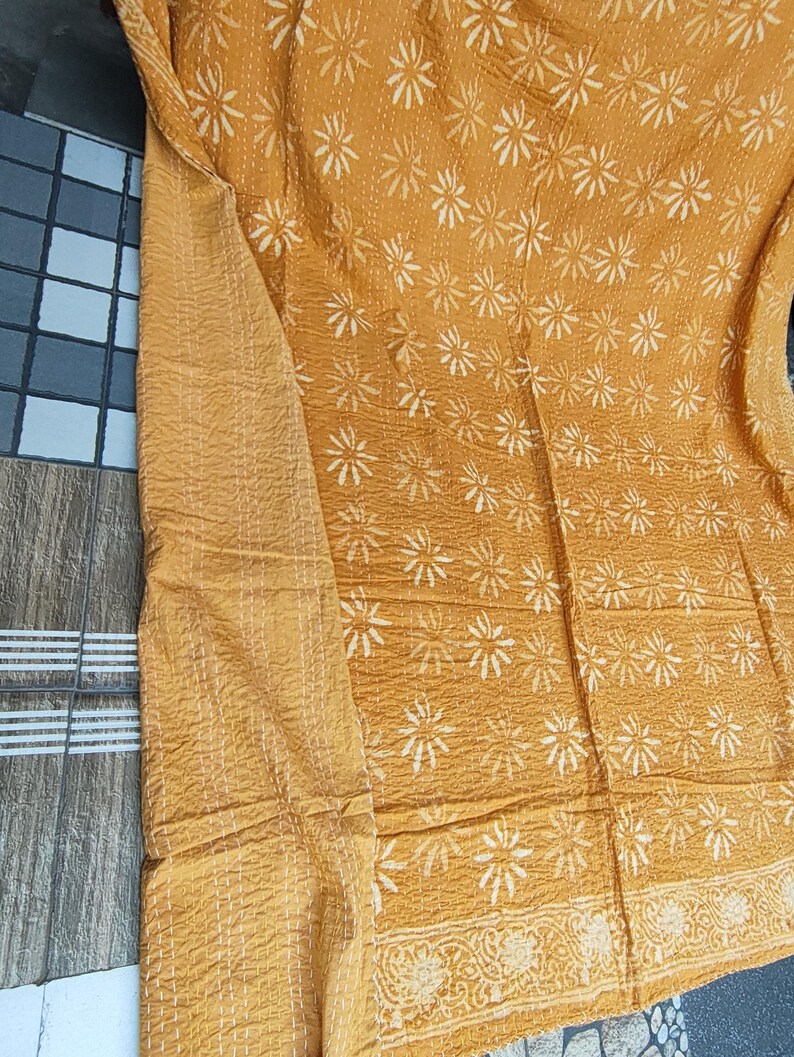 May include: Mustard-colored textile with a repeating white floral pattern. The fabric has a quilted texture with visible stitching. A detailed border with floral designs is visible along one edge. The textile is likely a blanket or throw.