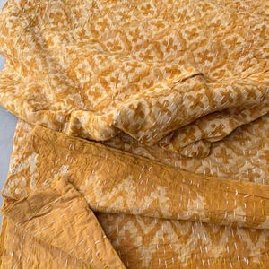 Op de afbeelding: Een gevouwen, geel en wit patroon quilt met zichtbare stiksels. De quilt heeft een herhalend geometrisch ontwerp in tinten geel en wit, met witte stiksels. De stof lijkt zacht en getextureerd.