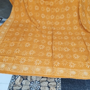 May include: Mustard yellow textile with a repeating white floral pattern. The edges feature a detailed border with floral and leaf designs. The fabric appears to be soft and possibly handmade, with visible stitching throughout the surface.