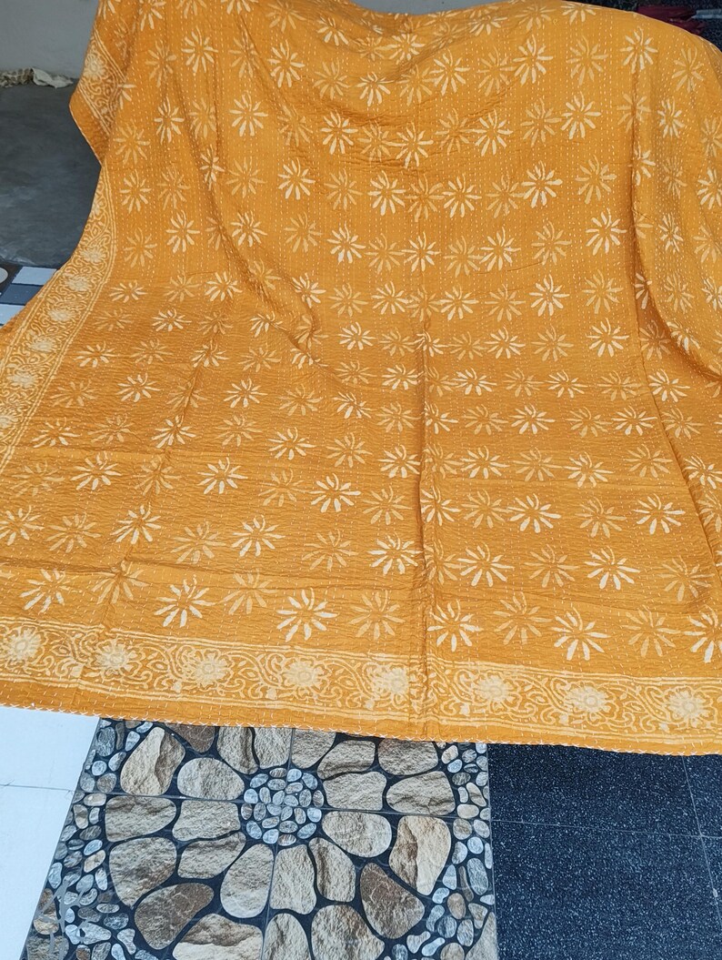 May include: Mustard yellow textile with a repeating white floral pattern. The edges feature a detailed border with floral and leaf designs. The fabric appears to be soft and possibly handmade, with visible stitching throughout the surface.