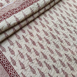 May include: Cream-colored quilted blanket with a repeating pattern of maroon leaf-like designs. The border features a maroon and white striped pattern with a zigzag design. The blanket is folded, showing its texture and design.