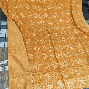 May include: Mustard-colored textile with a repeating white floral pattern. The fabric has a quilted appearance with visible stitching. A decorative border with a floral design is visible along the edge. The textile is folded, showing the front and the edge.