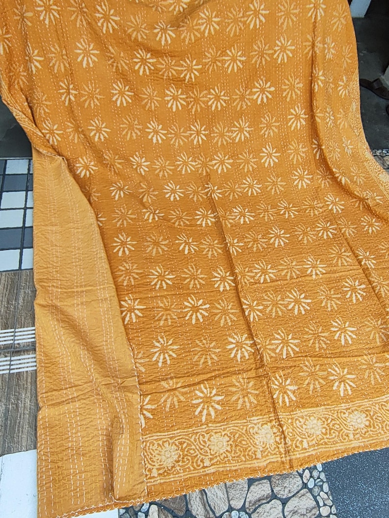 May include: Mustard-colored textile with a repeating white floral pattern. The fabric has a quilted appearance with visible stitching. A decorative border with a floral design is visible along the edge. The textile is folded, showing the front and the edge.