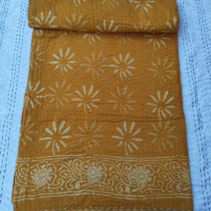 May include: Mustard yellow kantha quilt with a floral pattern and a decorative border. The quilt is folded, showcasing the intricate stitching and design. The background is a light blue fabric with a dotted pattern.