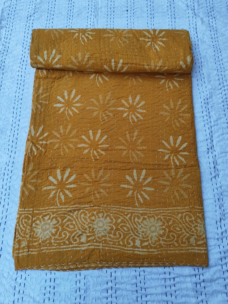 May include: Mustard yellow kantha quilt with a floral pattern and a decorative border. The quilt is folded, showcasing the intricate stitching and design. The background is a light blue fabric with a dotted pattern.