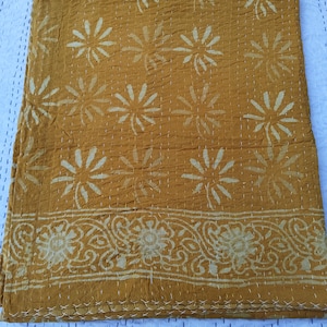 May include: Mustard-colored quilt with a floral pattern and decorative border. The quilt features a repeating design of large, stylized flowers and a detailed, scrolling border. White stitching is visible throughout the quilt, adding texture and detail to the design.