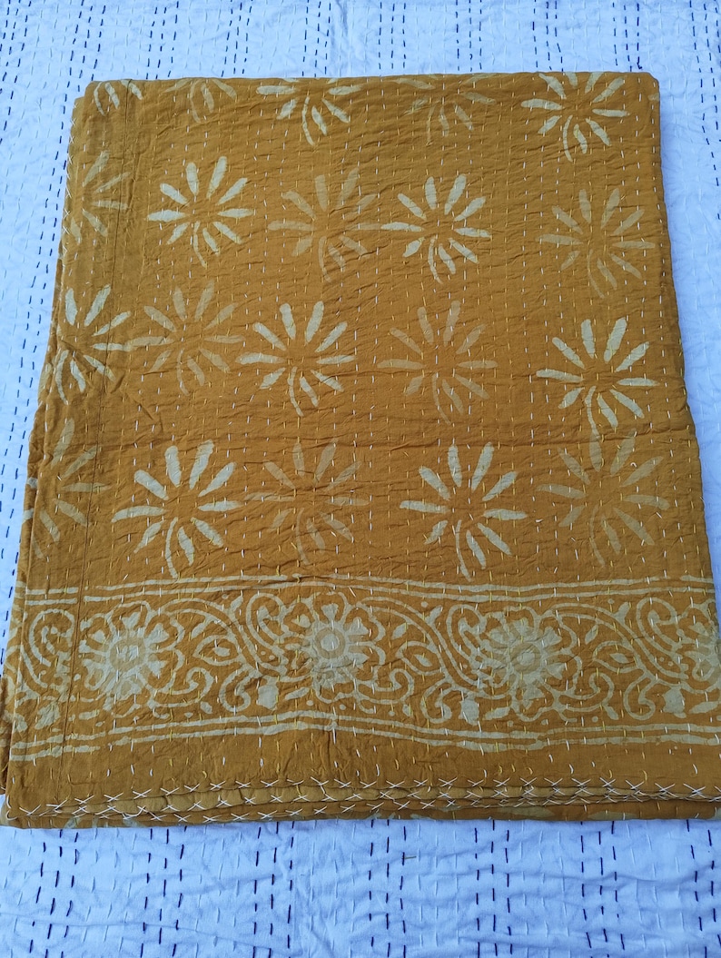 May include: Mustard-colored quilt with a floral pattern and decorative border. The quilt features a repeating design of large, stylized flowers and a detailed, scrolling border. White stitching is visible throughout the quilt, adding texture and detail to the design.
