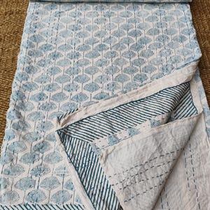 May include: A white and blue patterned quilt with a repeating fan-like design. The quilt is folded over, revealing a striped border and the stitched underside. The color palette is primarily white and light blue.