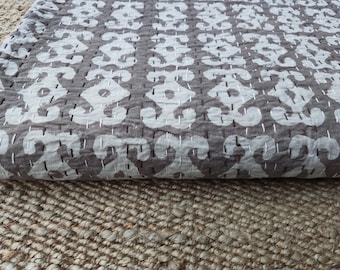 Puede incluir: Textil plegado con un patrón blanco repetido sobre un fondo gris. La tela está cosida con hilo blanco. El textil descansa sobre una superficie marrón texturizada. La estética general es rústica y artesanal, destacando el diseño de la tela.