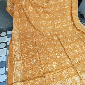 May include: Mustard-colored, floral-patterned quilt. The quilt features a repeating white flower design across the surface, with a detailed border. The fabric appears soft and textured, suitable for a bedspread or throw.