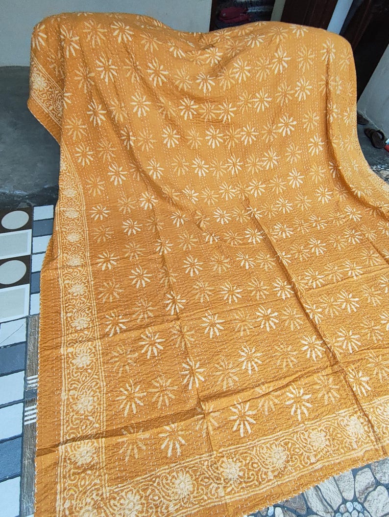 May include: Mustard-colored, floral-patterned quilt. The quilt features a repeating white flower design across the surface, with a detailed border. The fabric appears soft and textured, suitable for a bedspread or throw.