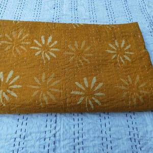 May include: Mustard-colored quilt with a floral pattern. The quilt features large, stylized white flowers evenly spaced across the surface. The stitching is visible, adding texture. The quilt is folded, showing its thickness and construction.