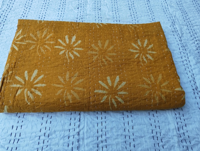 May include: Mustard-colored quilt with a floral pattern. The quilt features large, stylized white flowers evenly spaced across the surface. The stitching is visible, adding texture. The quilt is folded, showing its thickness and construction.