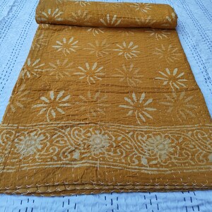 May include: Mustard yellow quilt with a floral pattern and decorative border. The quilt is folded and rolled, showcasing the intricate stitching and design. The background is a white surface with a stitched pattern.