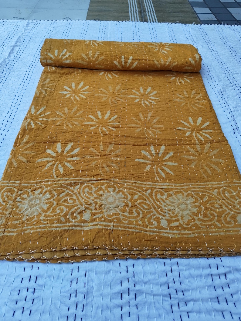 May include: Mustard yellow quilt with a floral pattern and decorative border. The quilt is folded and rolled, showcasing the intricate stitching and design. The background is a white surface with a stitched pattern.