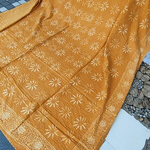 May include: Mustard-colored textile with a repeating white floral pattern. The edges are bordered with a detailed floral design. The fabric appears to be quilted, with visible stitching throughout. The textile is folded, showing its texture and design.