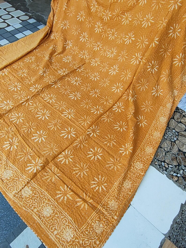 May include: Mustard-colored textile with a repeating white floral pattern. The edges are bordered with a detailed floral design. The fabric appears to be quilted, with visible stitching throughout. The textile is folded, showing its texture and design.