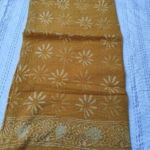 May include: Mustard-colored textile with a floral pattern. The fabric features a repeating design of stylized flowers in a lighter shade, with a decorative border along one edge. The textile is likely a throw or a decorative piece.