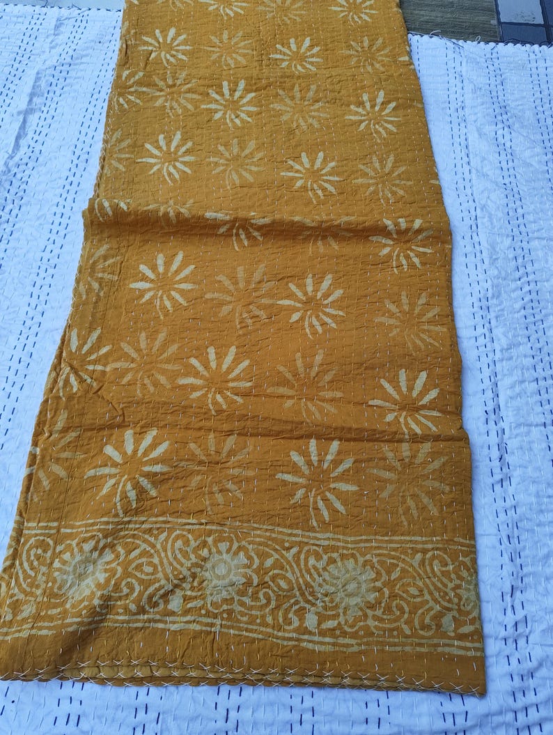 May include: Mustard-colored textile with a floral pattern. The fabric features a repeating design of stylized flowers in a lighter shade, with a decorative border along one edge. The textile is likely a throw or a decorative piece.