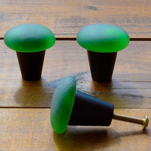Può includere: Quattro pomelli per armadi in vetro verde con basi nere. I pomelli sono rotondi e hanno una superficie leggermente ruvida. Sono disposti su una superficie in legno.