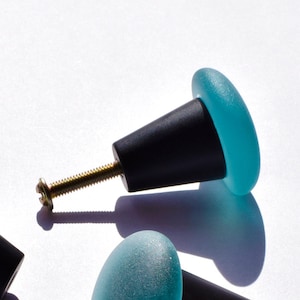 Puede incluir: Cinco pomos de armario de vidrio azul verdoso con bases de metal negro. Los pomos tienen un acabado de vidrio esmerilado.