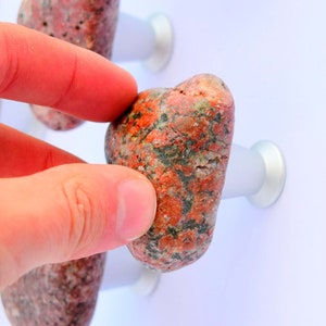 Könnte beinhalten: Nahaufnahme einer Hand, die einen roten und grauen Stein-Knopf hält. Der Knopf ist an einer weißen Wand mit einem silbernen Sockel befestigt. An der Wand befinden sich weitere ähnliche Knöpfe.