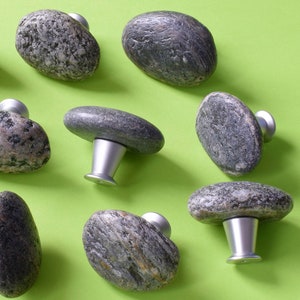 May include: Eleven stone cabinet knobs are displayed against a bright green backdrop. Each knob has a natural grey stone top and a silver-coloured metal base. The knobs' organic shapes and textures are highlighted in this arrangement.
