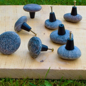 Könnte beinhalten: Eine Sammlung von Stein-Schrankknöpfen mit schwarzen Basen und Messingschrauben, angeordnet auf einem hellen Holzbrett. Die Knöpfe bestehen aus grau-rosa gesprenkelten Steinen und bieten eine natürliche, rustikale Ästhetik. Ein einzigartiger Einrichtungsgegenstand.