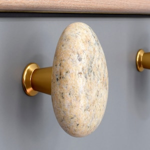 May include: Two cabinet knobs made of natural stone with a gold metal base. The knobs are shaped like rounded pebbles. One knob is a light beige colour with brown speckles, and the other knob is a pink and grey colour with black speckles.