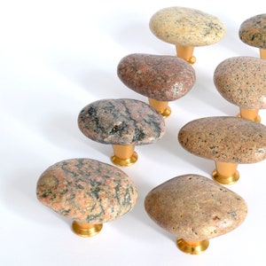 Könnte beinhalten: Acht natürliche Steinknaufgriffe mit goldener Oberfläche. Die Knäufe sind flach, oval geformt und in verschiedenen Brauntönen, Grautönen und Rosatönen gehalten.