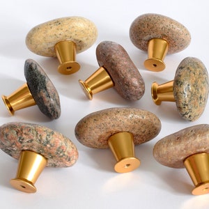 Könnte beinhalten: Acht natürliche Steinknaufgriffe mit goldenen Metallsockeln. Die Knäufe sind in Form von flachen, ovalen Steinen mit einer Vielzahl von braunen, grauen und weißen Mustern geformt.