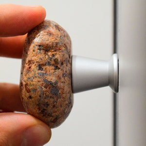 Könnte beinhalten: Eine Hand hält einen braunen und grauen Steintürknopf mit einer silbernen Basis.