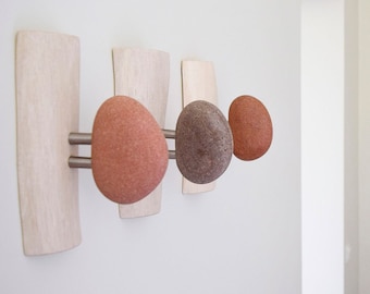 May include: A wall-mounted coat rack featuring four natural stone knobs in shades of red and brown. The knobs are mounted on brushed metal rods and set against light wood panels, creating a modern, organic design.