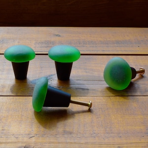 May include: Four green glass cabinet knobs with black bases. The knobs are round and have a slightly textured surface. One knob is laying on its side with a gold screw visible.