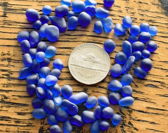 May include: A close-up of a pile of blue sea glass pieces arranged in a circle around a silver US nickel coin. The coin features the words "E Pluribus Unum" and "Five Cents".