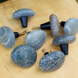 May include: Eight grey stone cabinet knobs with black metal hardware. The knobs are various shapes and sizes, with a natural, rough texture.
