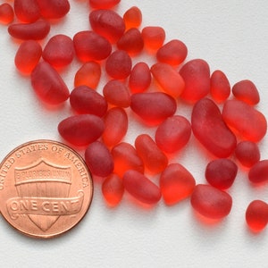 Könnte beinhalten: Eine Sammlung kleiner, glatter, roter Glassteine. Die Steine sind auf einer weißen Oberfläche verstreut. Ein Cent-Stück ist zur Größenbestimmung enthalten.