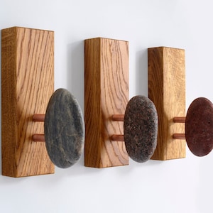 May include: Three wooden wall hooks with natural stone knobs. The hooks are made of light brown wood with a grain pattern. The stones are different colors and shapes. The stones are attached to the wood with copper screws.