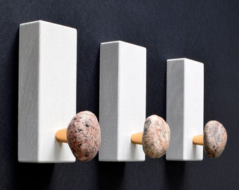 May include: Three white rectangular wall hooks with natural stone knobs. The hooks are mounted on a black wall.