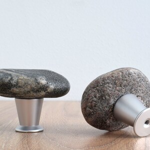 以下が含まれることがあります： 銀色の金属製のベースが付いた灰色の石製のキャビネットノブ2個。ノブは平らで丸みを帯びた石の形をしています。