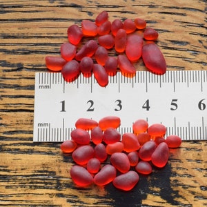 May include: A close-up of a pile of small, red glass beads measured against a ruler. The ruler is marked in millimeters and shows measurements from 1 to 8.